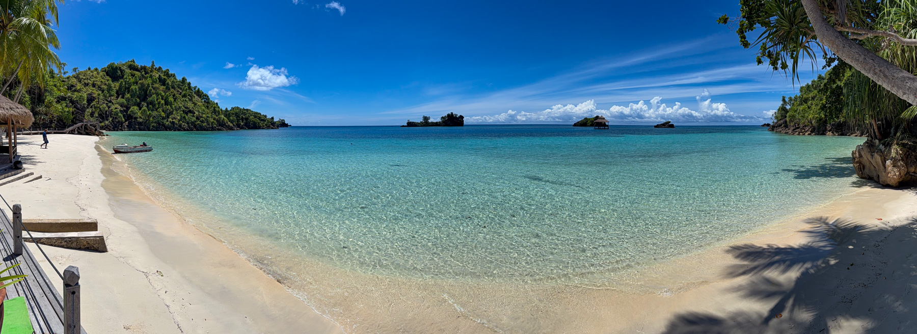 A panorama of the swimming bay, flat water shading from clear to deep blue, a skiff pulled up on the sand at the left and small forested islets on the horizon