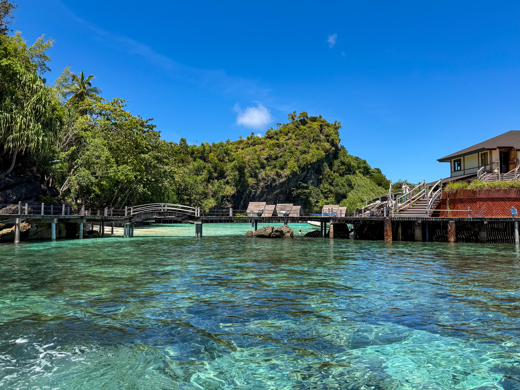 The jetty seen from the water, an arched timber bridge in its span, sun loungers under thatched shades along the deck and the main building above the rocks on the right