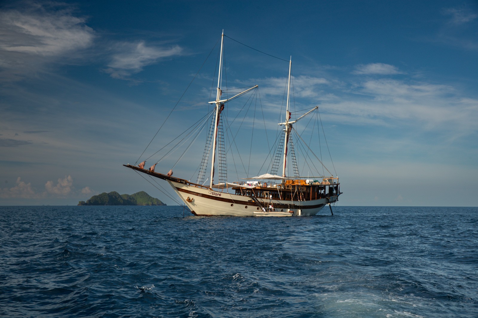 A two-masted wooden phinisi lying at anchor on flat water, an island low on the horizon behind