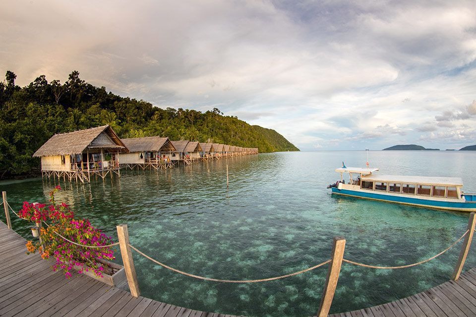 Over-water bungalows on stilts above clear shallows, a jetty and a boat beyond them, forested hills behind