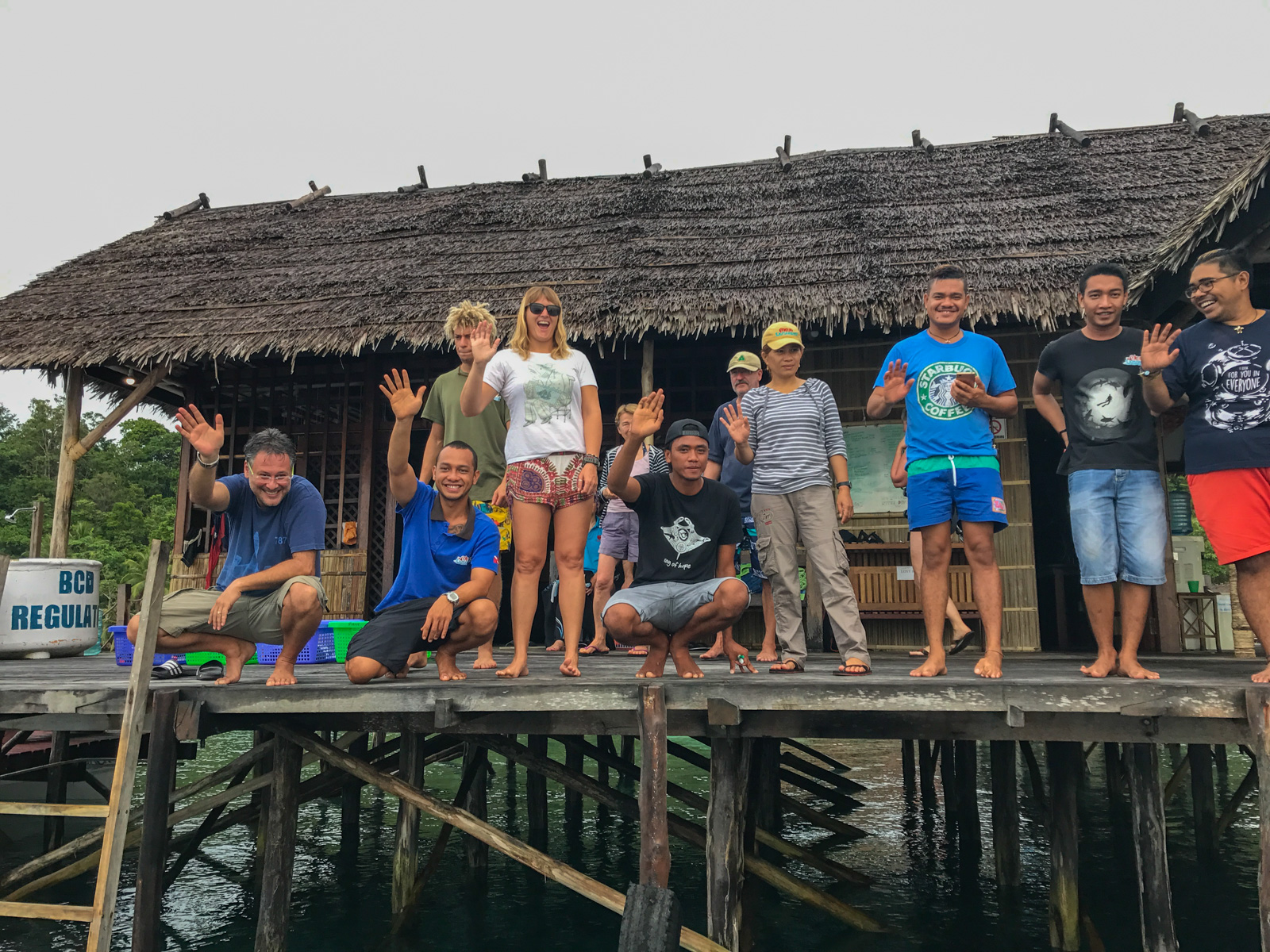 The dive team and guests on the jetty outside the thatched dive centre, most of them waving.