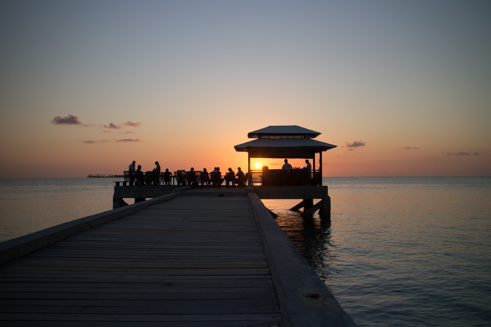 A long jetty out to a thatched shelter, silhouetted figures at the end against a low sun